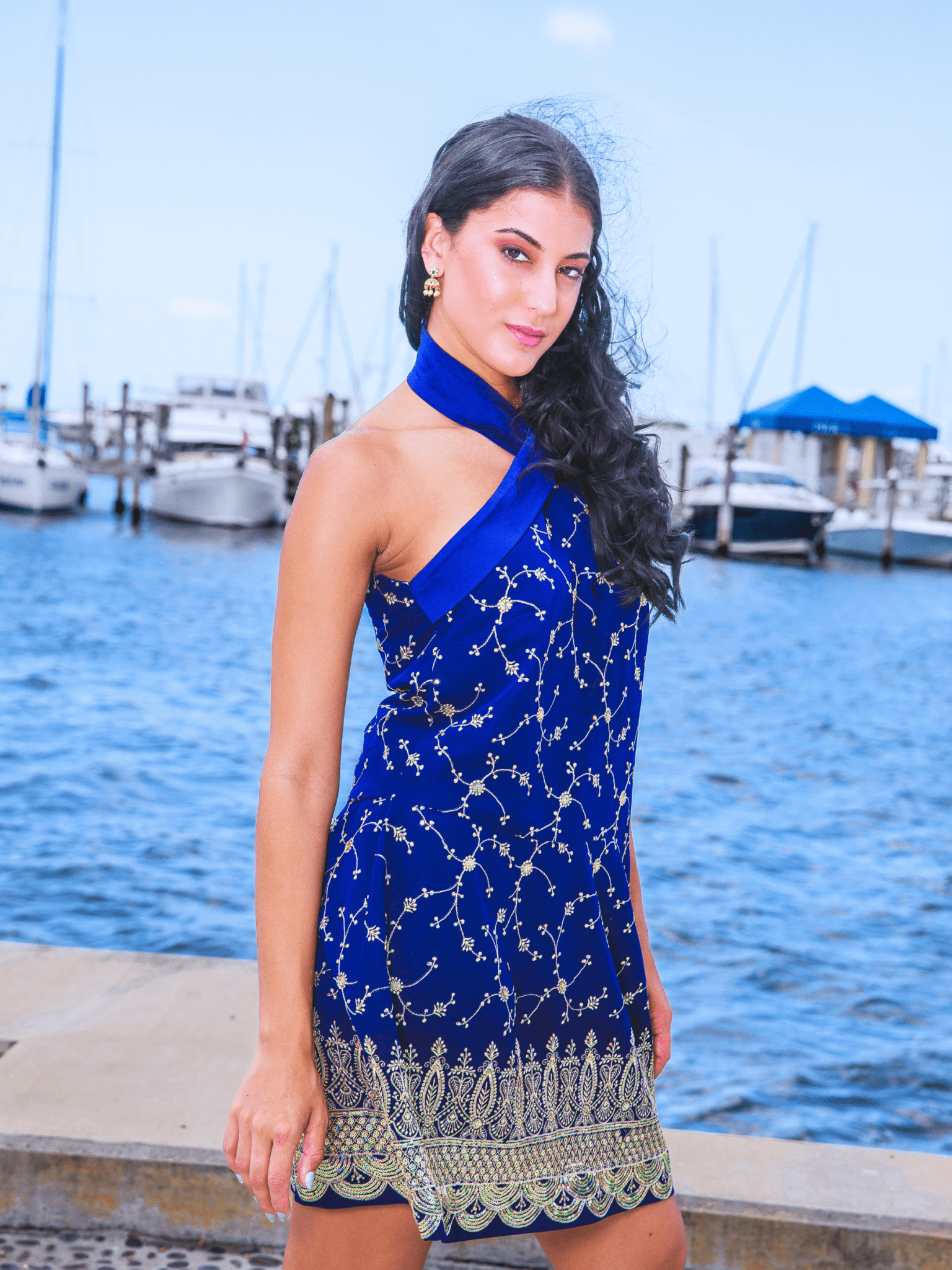 Woman in a blue dress with star patterns standing by a waterfront with boats.