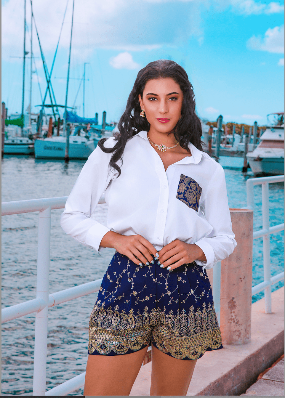 Woman in a white shirt and patterned shorts standing by a waterfront with boats in the background