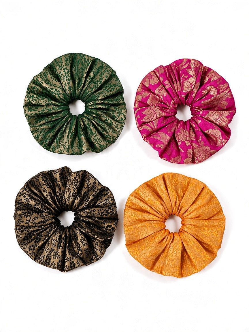 Four fabric hair scrunchies arranged in a 2x2 grid on a white background. The top-left scrunchie is dark green with a metallic gold pattern, the top-right is bright pink with a gold floral pattern, the bottom-left is black with a gold pattern, and the bottom-right is a solid golden orange.
