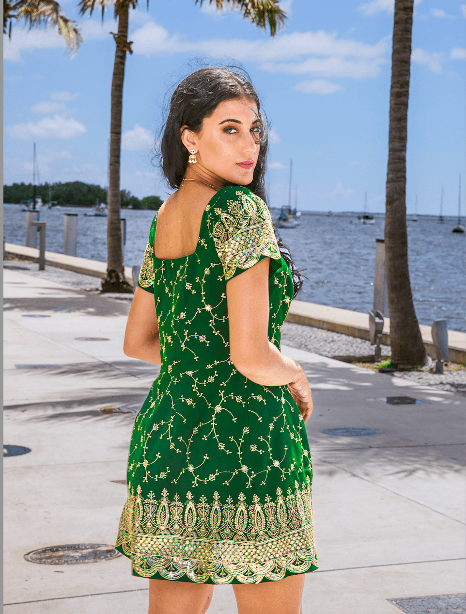 Woman in a green dress with gold patterns standing by a waterfront with palm trees and boats.