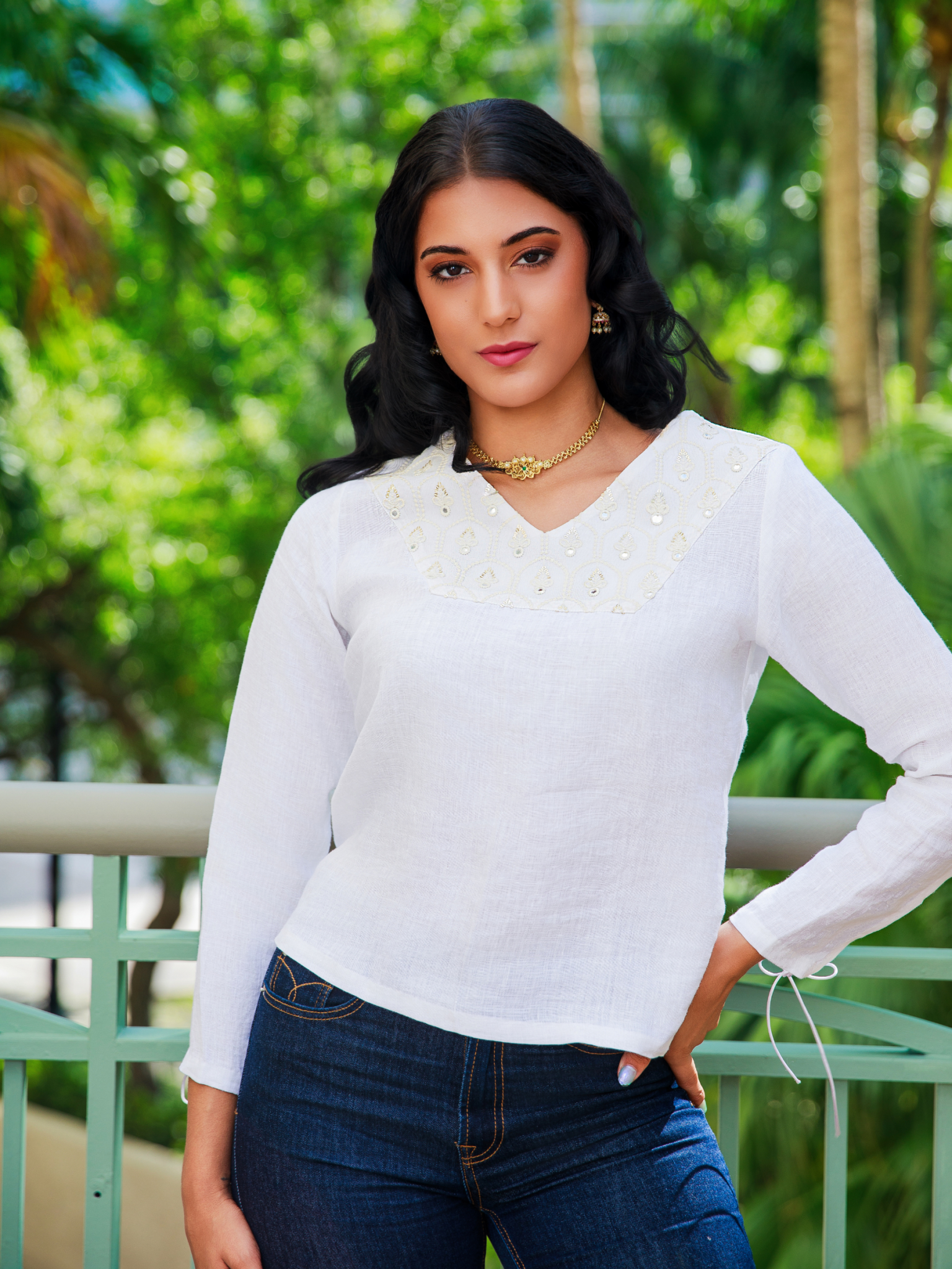 Woman wearing a white sweater and blue jeans standing outdoors with greenery in the background
