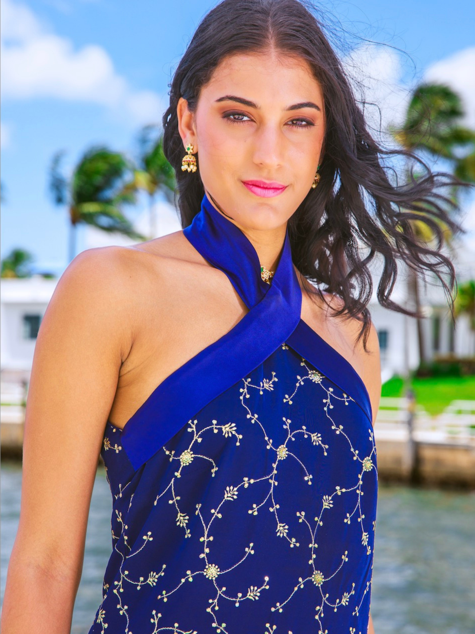 Woman wearing a blue dress with floral patterns by a body of water with palm trees in the background