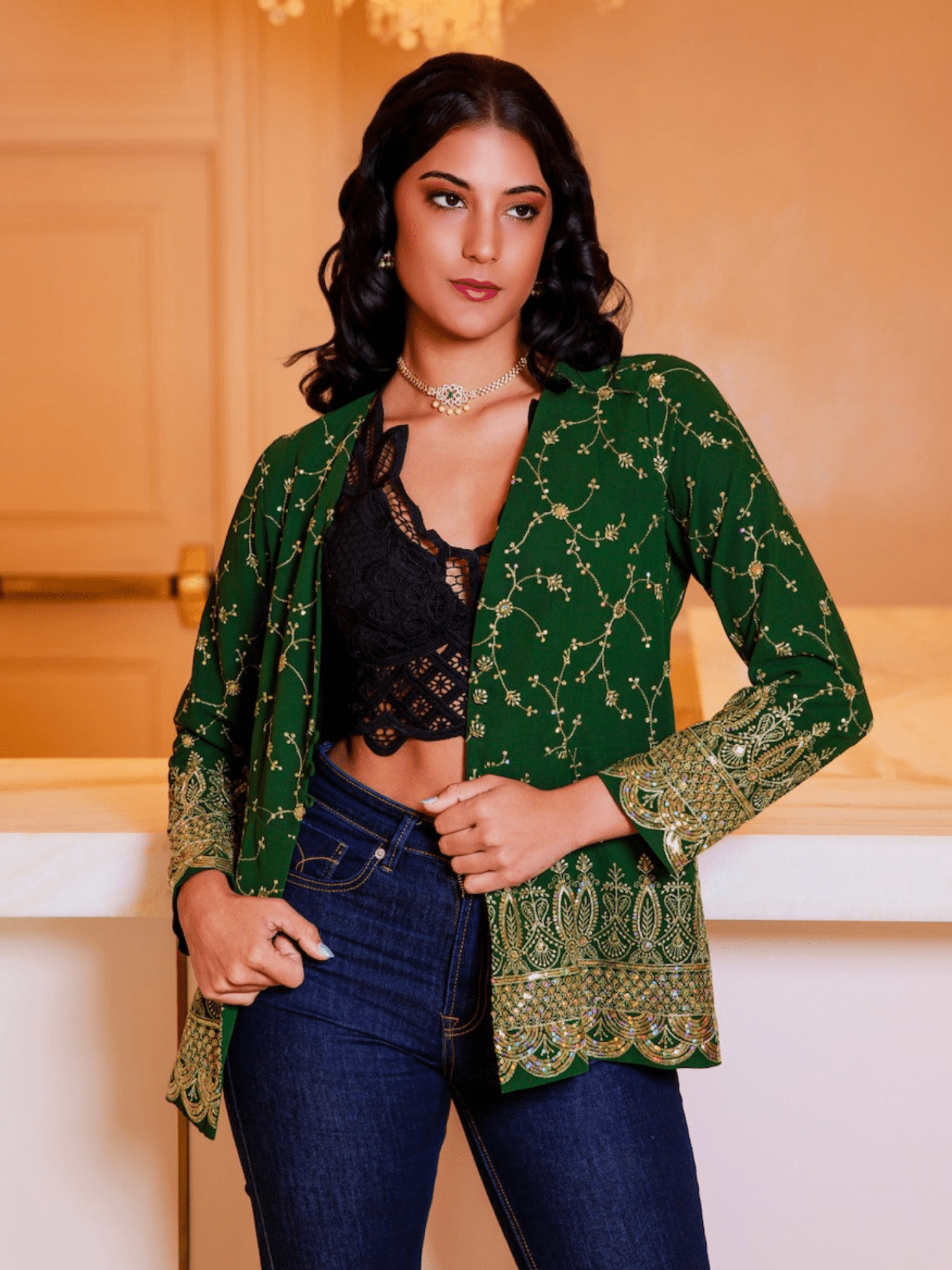 Woman wearing a green embroidered jacket over a black top and blue jeans indoors.