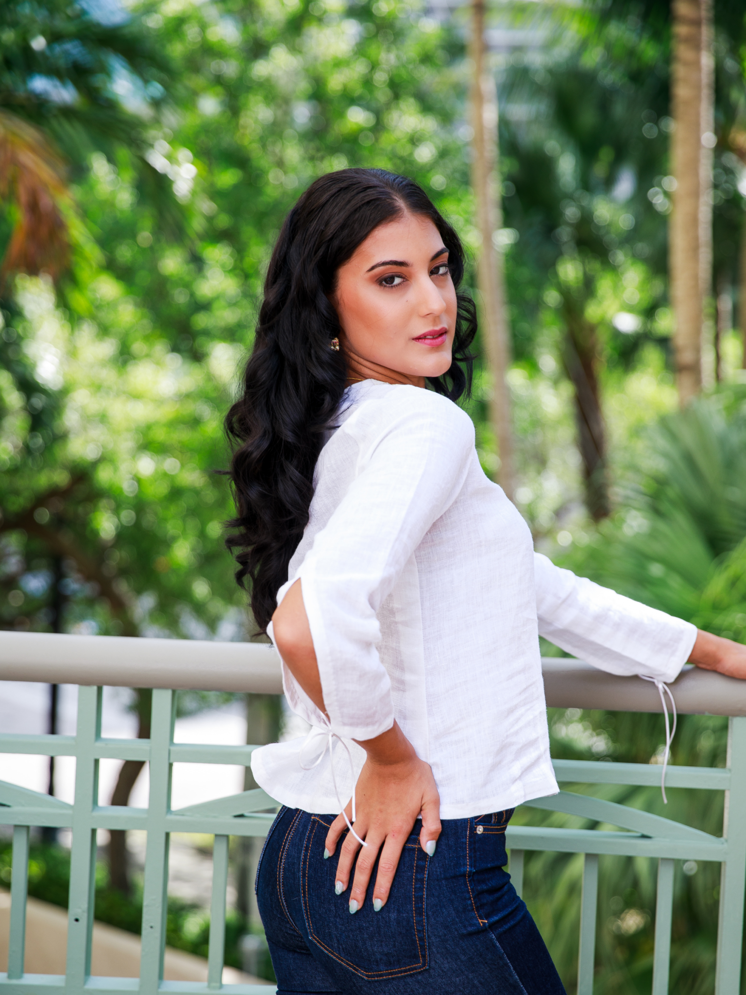 Woman in a white blouse and blue jeans standing outdoors with greenery in the background