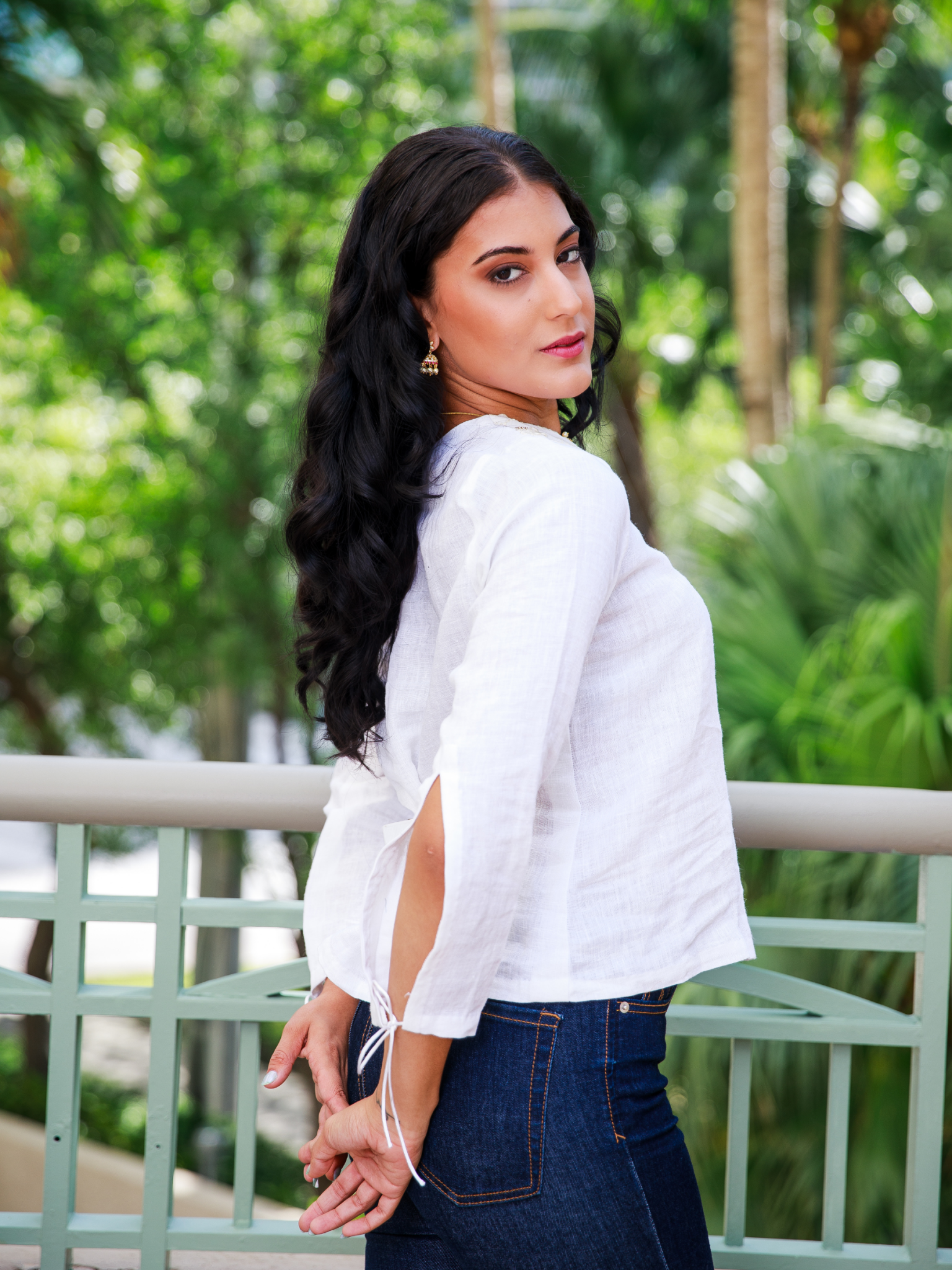 Woman wearing a white blouse with a cut-out design, standing outdoors with greenery in the background.