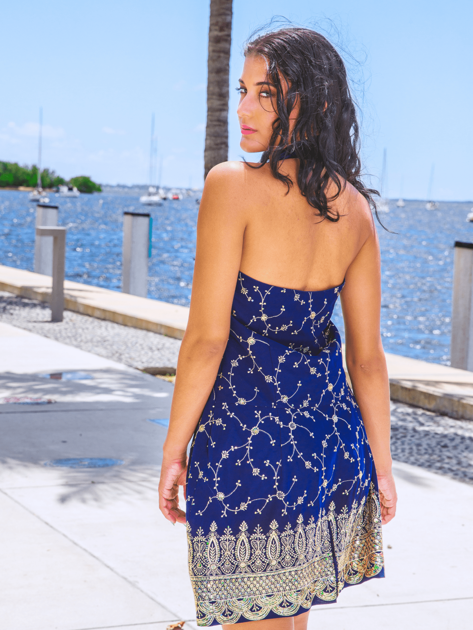 Woman in a blue dress with gold patterns standing by a waterfront.