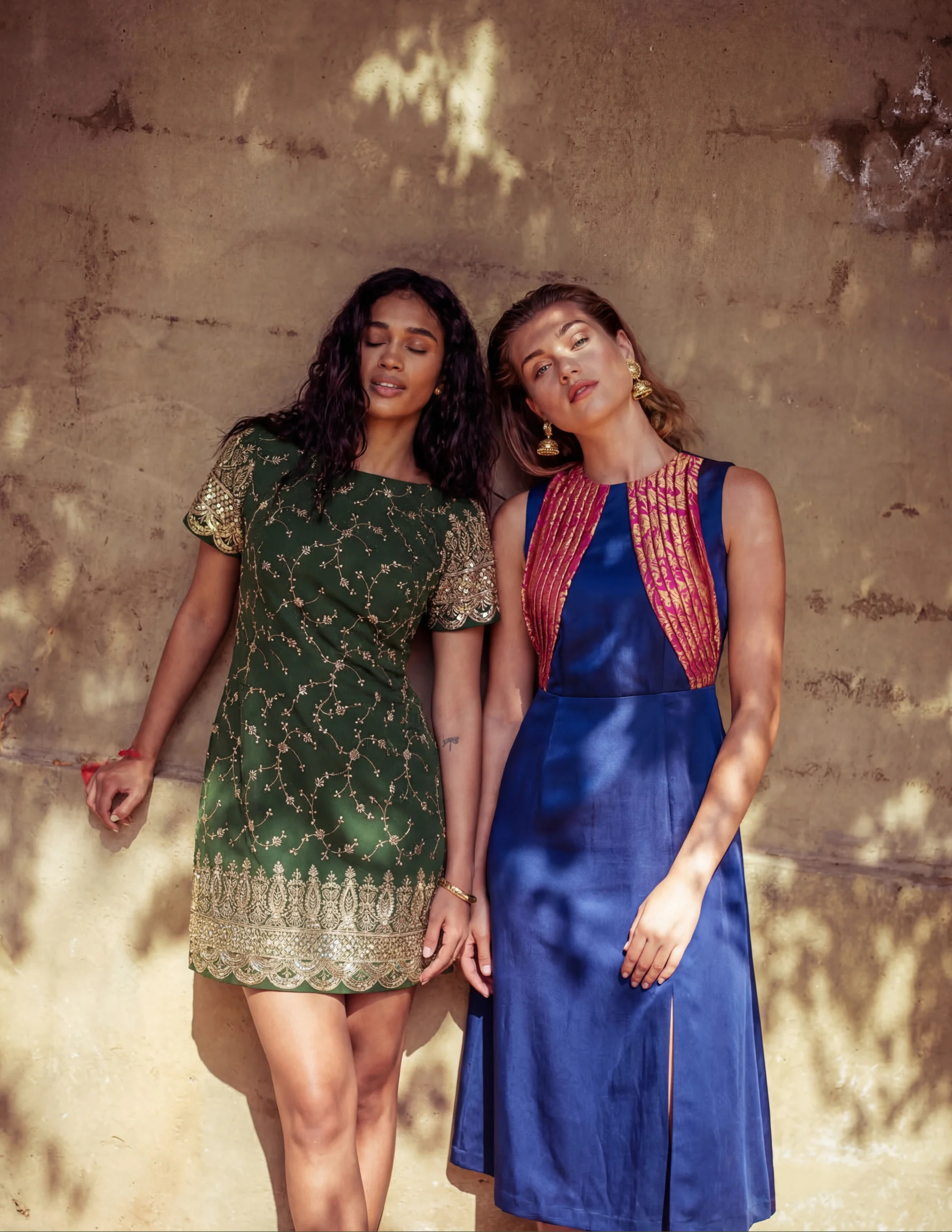 Two women in traditional outfits standing against a textured wall.