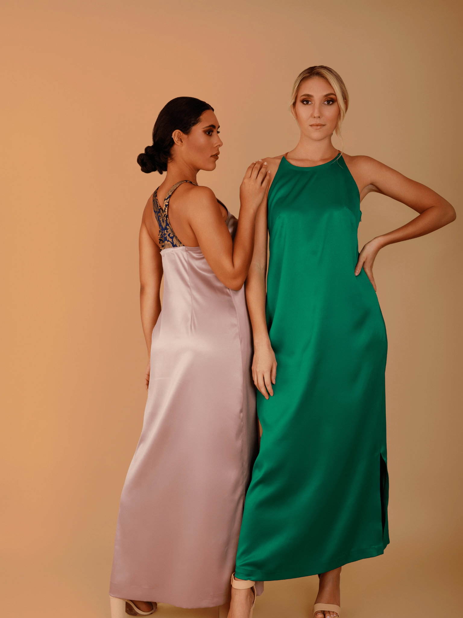 Two women wearing long dresses, one pink and one green, against a beige background.