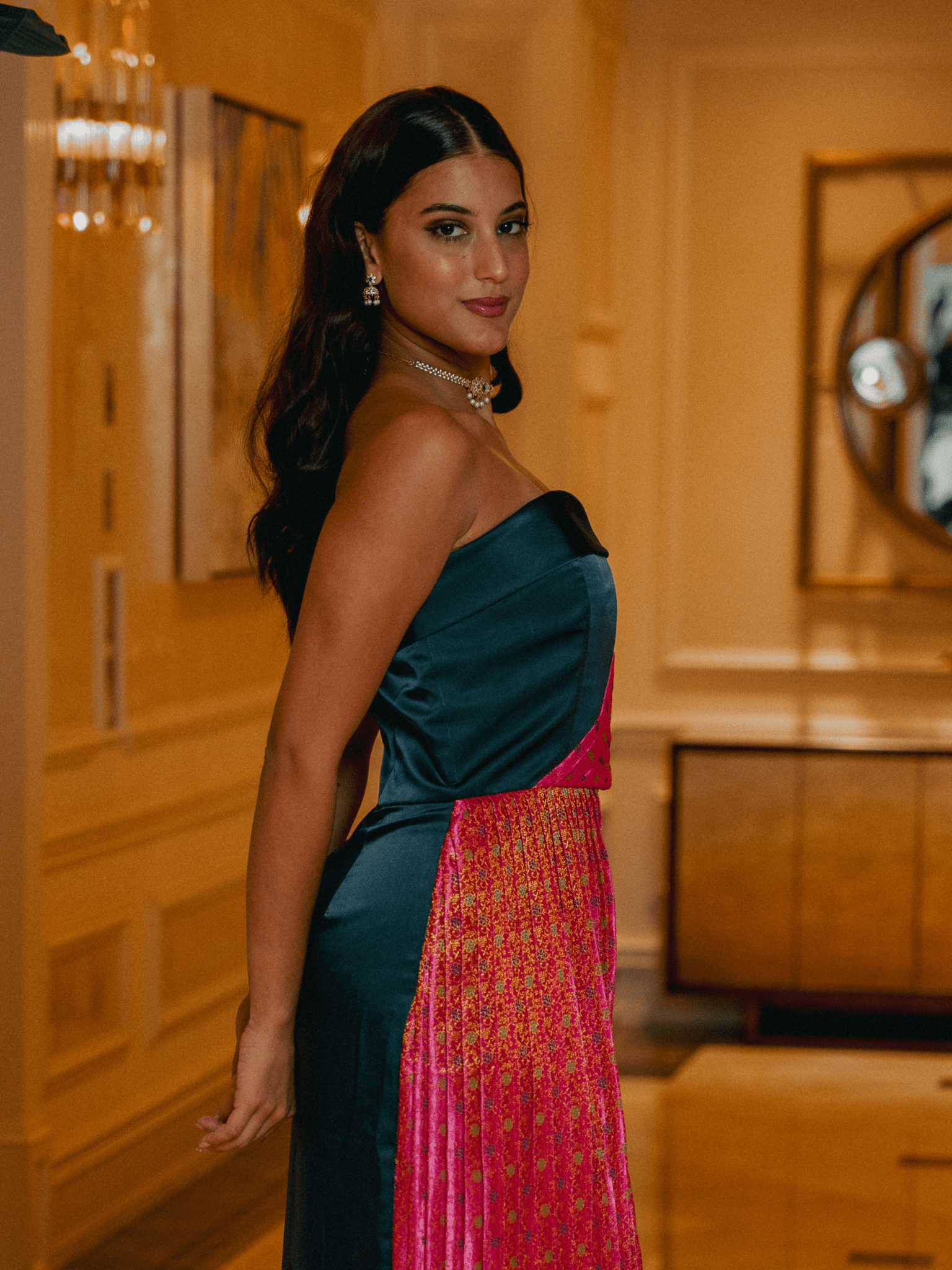 Woman in a strapless teal and pink dress standing in an elegant room with gold walls.