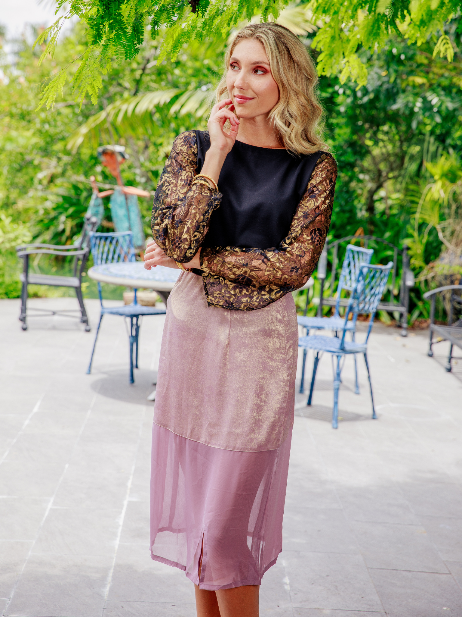 Woman in a black top and pink skirt standing outdoors with greenery in the background