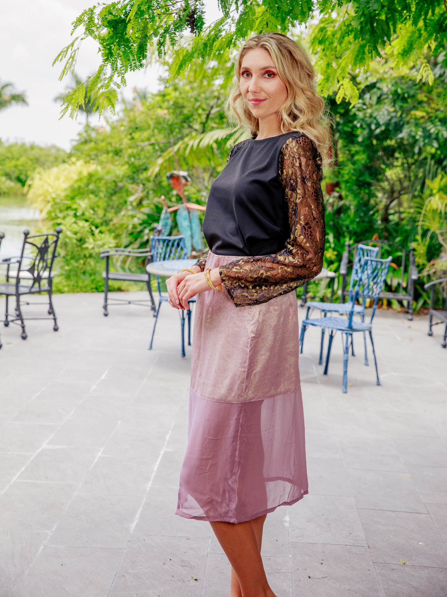 Woman in a black top and pink skirt standing outdoors with greenery in the background