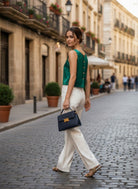 A woman walking on a cobblestone street wearing a sleeveless emerald green top with gold buttons, white wide-leg trousers, and carrying a navy blue handbag with gold hardware.