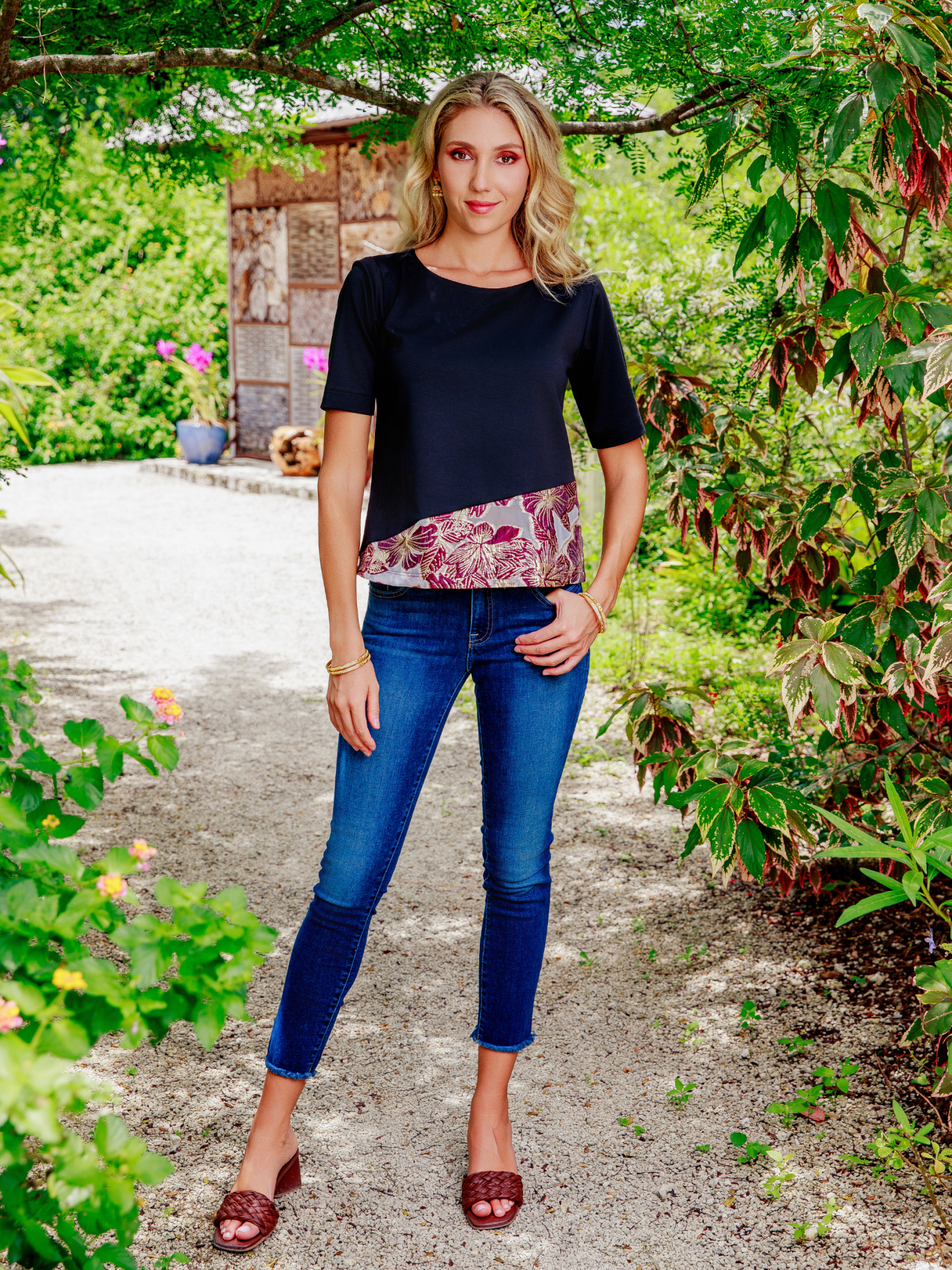 Woman standing outdoors in a garden wearing a black top with a floral pattern and blue jeans.