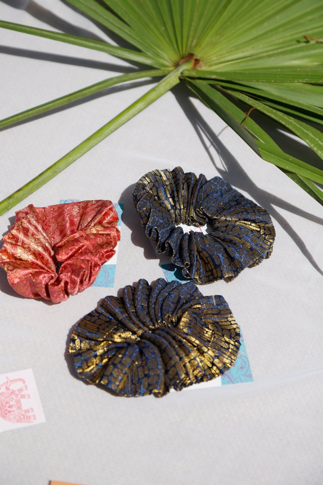 Three artisanal scrunchies arranged on a surface with a tropical leaf background. The scrunchies are in shades of red, blue, and gold, featuring a textured and decorative pattern.