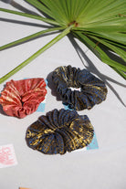 Three artisanal scrunchies arranged on a surface with a tropical leaf background. The scrunchies are in shades of red, blue, and gold, featuring a textured and decorative pattern.